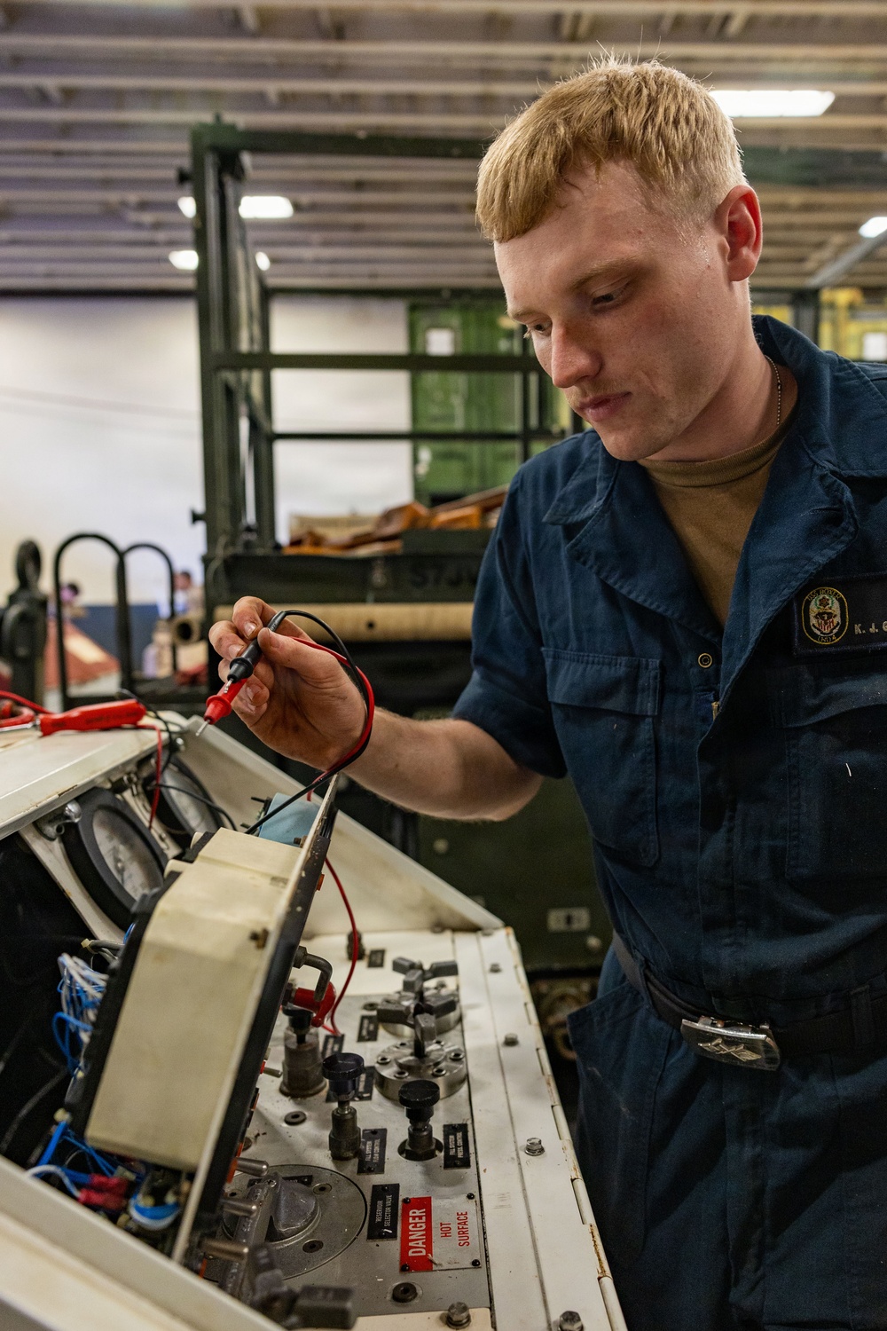 A young male sailor in U.S. Navy blue coveralls is using a multimeter with red and black test probes on an open equipment panel. The panel contains various gauges, switches, knobs, and wiring, with a visible 'DANGER HOT SURFACE' warning label. The setting appears to be a military maintenance bay or hangar facility.
