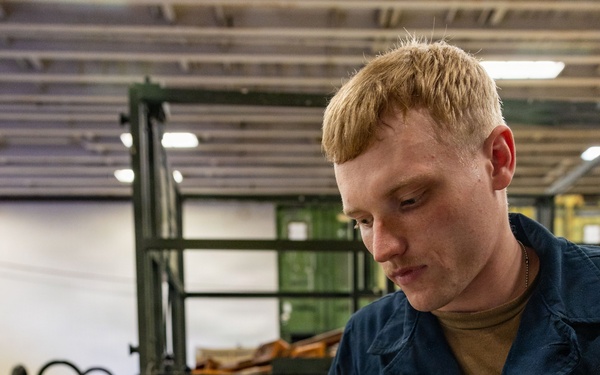 A young male sailor in U.S. Navy blue coveralls is using a multimeter with red and black test probes on an open equipment panel. The panel contains various gauges, switches, knobs, and wiring, with a visible 'DANGER HOT SURFACE' warning label. The setting appears to be a military maintenance bay or hangar facility.