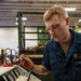 A young male sailor in U.S. Navy blue coveralls is using a multimeter with red and black test probes on an open equipment panel. The panel contains various gauges, switches, knobs, and wiring, with a visible 'DANGER HOT SURFACE' warning label. The setting appears to be a military maintenance bay or hangar facility.
