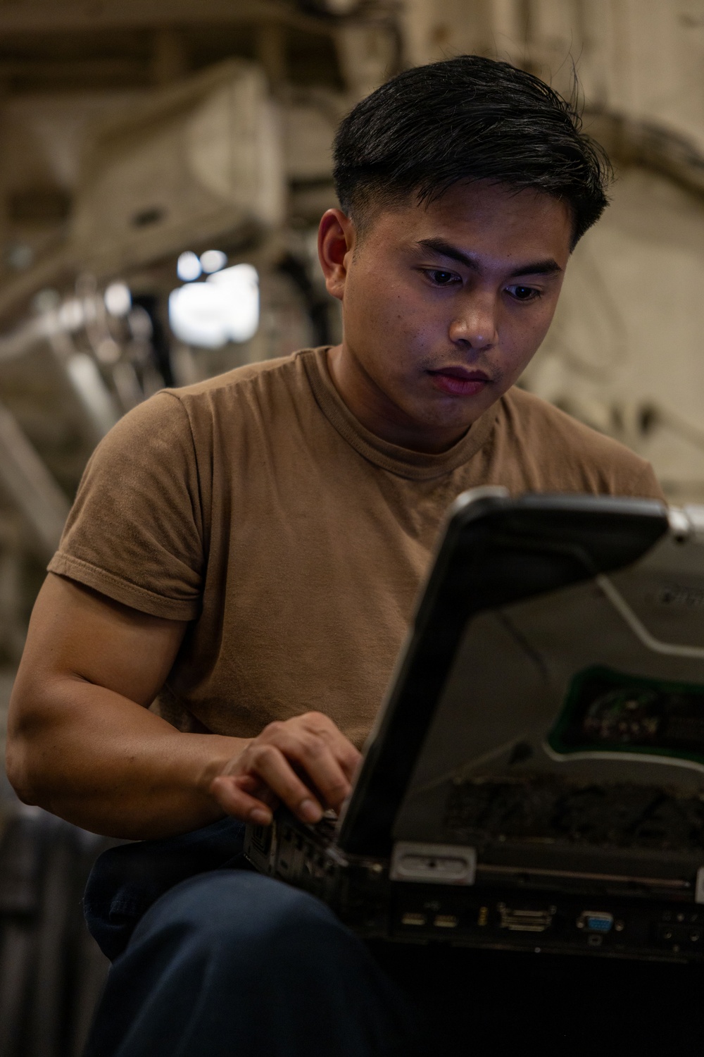 A young man wearing a tan/brown t-shirt is focused on operating a ruggedized military-style laptop computer. He appears to be working in an industrial or military environment, possibly inside a large aircraft or vehicle based on the structural elements visible in the background. His expression is concentrated and attentive to the screen.