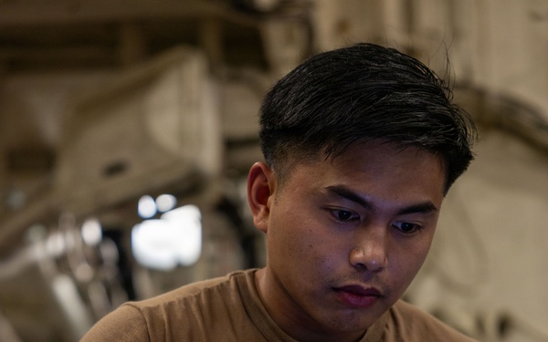 A young man wearing a tan/brown t-shirt is focused on operating a ruggedized military-style laptop computer. He appears to be working in an industrial or military environment, possibly inside a large aircraft or vehicle based on the structural elements visible in the background. His expression is concentrated and attentive to the screen.