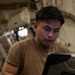 A young man wearing a tan/brown t-shirt is focused on operating a ruggedized military-style laptop computer. He appears to be working in an industrial or military environment, possibly inside a large aircraft or vehicle based on the structural elements visible in the background. His expression is concentrated and attentive to the screen.