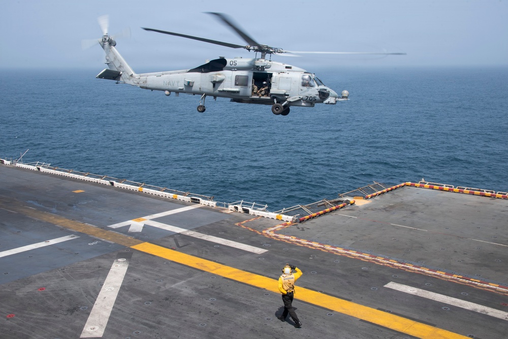 A U.S. Navy helicopter, marked with the number '05' and '705', is hovering over the flight deck of a naval vessel while a deck crew member in a yellow jersey signals the aircraft. The scene takes place at sea with calm, gray-blue ocean water visible in the background. This appears to be a routine flight deck operation, likely a landing or hover exercise.