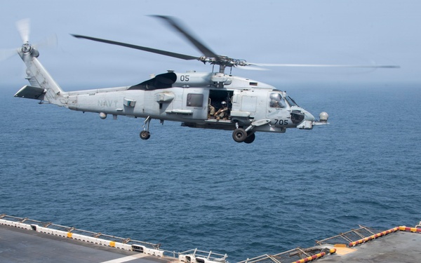 A U.S. Navy helicopter, marked with the number '05' and '705', is hovering over the flight deck of a naval vessel while a deck crew member in a yellow jersey signals the aircraft. The scene takes place at sea with calm, gray-blue ocean water visible in the background. This appears to be a routine flight deck operation, likely a landing or hover exercise.