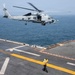 A U.S. Navy helicopter, marked with the number '05' and '705', is hovering over the flight deck of a naval vessel while a deck crew member in a yellow jersey signals the aircraft. The scene takes place at sea with calm, gray-blue ocean water visible in the background. This appears to be a routine flight deck operation, likely a landing or hover exercise.