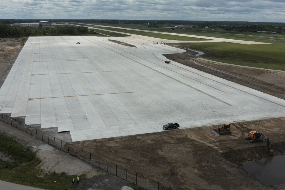 An aerial view of a large airport apron or taxiway under construction, featuring freshly laid concrete pavement slabs in a grid pattern. The site appears to be in a late stage of construction with surrounding earthwork still ongoing. Construction equipment, vehicles, and workers are visible around the perimeter of the paved area.