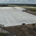 An aerial view of a large airport apron or taxiway under construction, featuring freshly laid concrete pavement slabs in a grid pattern. The site appears to be in a late stage of construction with surrounding earthwork still ongoing. Construction equipment, vehicles, and workers are visible around the perimeter of the paved area.