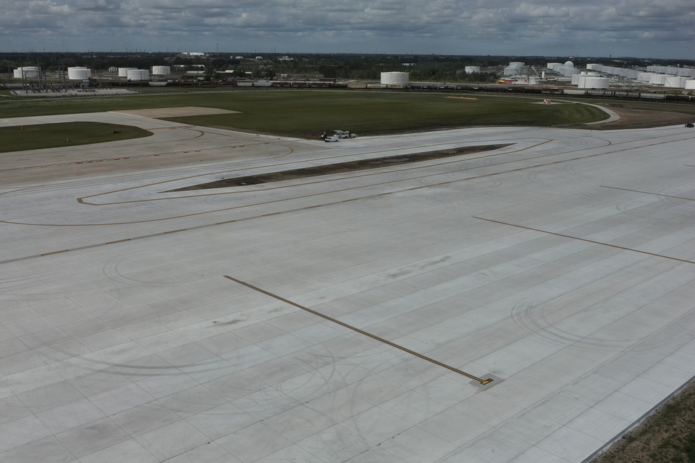 An aerial view of a large airport apron or tarmac area showing concrete pavement with visible tire marks and taxiway markings. The surrounding area includes grassy infields, a service vehicle, and industrial storage tanks visible in the background. The scene appears to be at a commercial or industrial airport located near a petroleum storage facility.