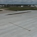 An aerial view of a large airport apron or tarmac area showing concrete pavement with visible tire marks and taxiway markings. The surrounding area includes grassy infields, a service vehicle, and industrial storage tanks visible in the background. The scene appears to be at a commercial or industrial airport located near a petroleum storage facility.