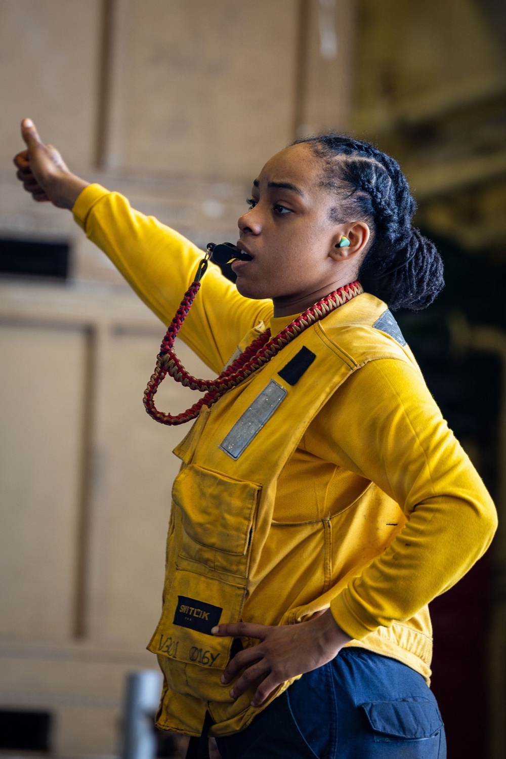A person wearing a yellow flight deck vest and yellow long-sleeve shirt is giving a thumbs-up hand signal while blowing a whistle. They are wearing hearing protection (green earplugs) and have a red-and-tan braided lanyard around their neck attached to the whistle. The setting appears to be an indoor industrial or military environment, consistent with a naval vessel hangar bay.