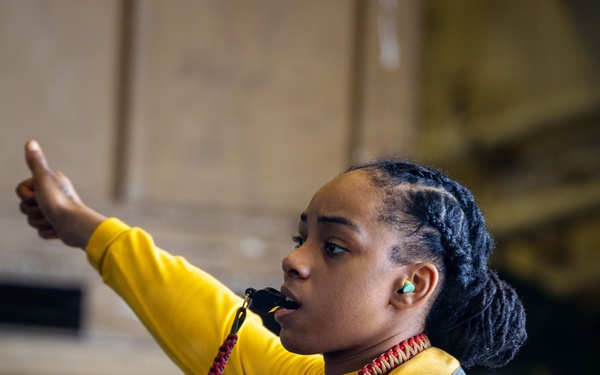 A person wearing a yellow flight deck vest and yellow long-sleeve shirt is giving a thumbs-up hand signal while blowing a whistle. They are wearing hearing protection (green earplugs) and have a red-and-tan braided lanyard around their neck attached to the whistle. The setting appears to be an indoor industrial or military environment, consistent with a naval vessel hangar bay.