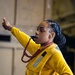 A person wearing a yellow flight deck vest and yellow long-sleeve shirt is giving a thumbs-up hand signal while blowing a whistle. They are wearing hearing protection (green earplugs) and have a red-and-tan braided lanyard around their neck attached to the whistle. The setting appears to be an indoor industrial or military environment, consistent with a naval vessel hangar bay.