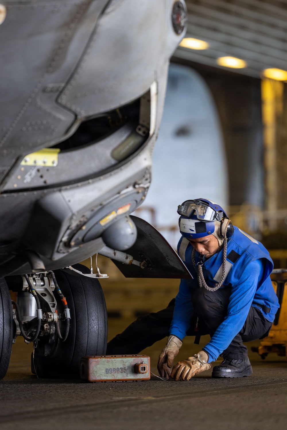 A military aircraft maintainer in a blue flight deck jersey is crouching beneath the nose landing gear of a military jet aircraft, performing maintenance work. The setting appears to be an aircraft carrier hangar bay, indicated by the overhead lighting and structural elements visible in the background. The technician is working near a small equipment box labeled 'D9B035' on the hangar deck floor.
