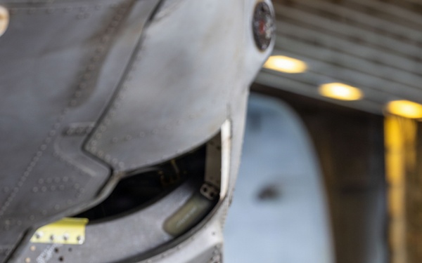 A military aircraft maintainer in a blue flight deck jersey is crouching beneath the nose landing gear of a military jet aircraft, performing maintenance work. The setting appears to be an aircraft carrier hangar bay, indicated by the overhead lighting and structural elements visible in the background. The technician is working near a small equipment box labeled 'D9B035' on the hangar deck floor.