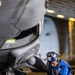 A military aircraft maintainer in a blue flight deck jersey is crouching beneath the nose landing gear of a military jet aircraft, performing maintenance work. The setting appears to be an aircraft carrier hangar bay, indicated by the overhead lighting and structural elements visible in the background. The technician is working near a small equipment box labeled 'D9B035' on the hangar deck floor.