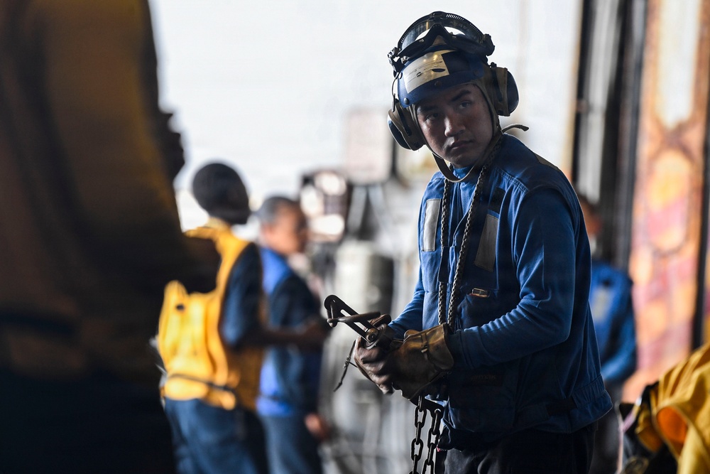 A military or naval deck crew member in the foreground is wearing a blue uniform, helmet with goggles, hearing protection, and gloves while holding a chain and hook assembly. The setting appears to be an aircraft carrier flight deck or hangar bay, with additional crew members visible in the background wearing yellow and blue vests. The image captures what appears to be routine aircraft handling or tie-down operations.