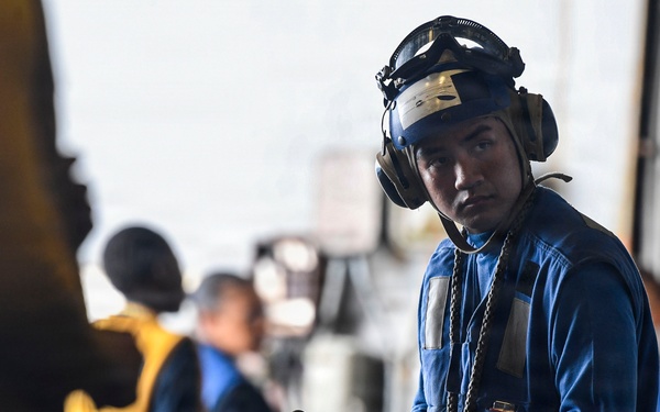 A military or naval deck crew member in the foreground is wearing a blue uniform, helmet with goggles, hearing protection, and gloves while holding a chain and hook assembly. The setting appears to be an aircraft carrier flight deck or hangar bay, with additional crew members visible in the background wearing yellow and blue vests. The image captures what appears to be routine aircraft handling or tie-down operations.