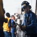 A military or naval deck crew member in the foreground is wearing a blue uniform, helmet with goggles, hearing protection, and gloves while holding a chain and hook assembly. The setting appears to be an aircraft carrier flight deck or hangar bay, with additional crew members visible in the background wearing yellow and blue vests. The image captures what appears to be routine aircraft handling or tie-down operations.