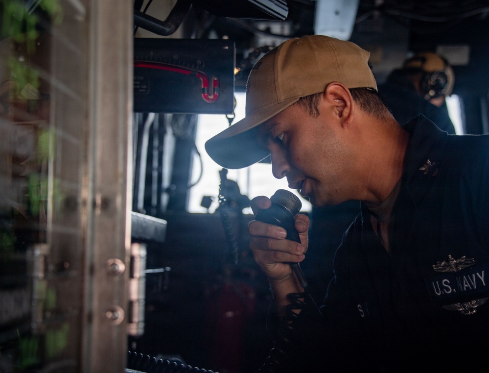 A U.S. Navy sailor is shown speaking into a handheld radio or intercom device aboard what appears to be a naval vessel's bridge or command area. The sailor is wearing a tan baseball cap and dark navy uniform with visible 'U.S. NAVY' text and qualification insignia. The setting features various shipboard electronic equipment and controls in the background.