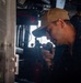 A U.S. Navy sailor is shown speaking into a handheld radio or intercom device aboard what appears to be a naval vessel's bridge or command area. The sailor is wearing a tan baseball cap and dark navy uniform with visible 'U.S. NAVY' text and qualification insignia. The setting features various shipboard electronic equipment and controls in the background.