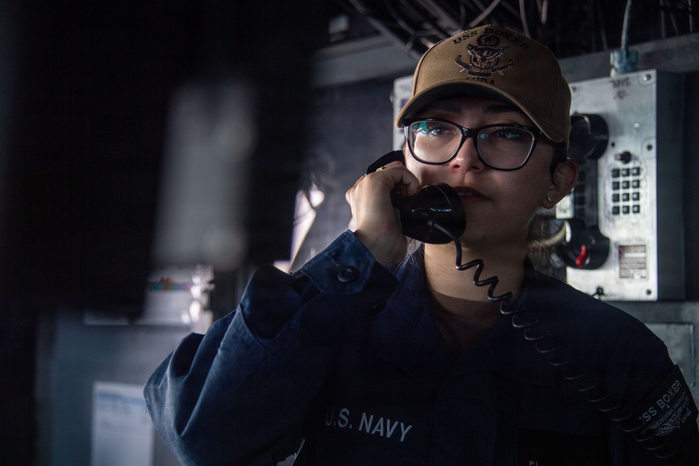 A U.S. Navy sailor is photographed in a dimly lit shipboard environment while speaking on a sound-powered or internal communications telephone. She is wearing a navy blue uniform with 'U.S. NAVY' visible on the sleeve and a tan ball cap bearing a ship's insignia. The image has a dramatic, low-key lighting style consistent with professional military photography.