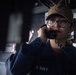 A U.S. Navy sailor is photographed in a dimly lit shipboard environment while speaking on a sound-powered or internal communications telephone. She is wearing a navy blue uniform with 'U.S. NAVY' visible on the sleeve and a tan ball cap bearing a ship's insignia. The image has a dramatic, low-key lighting style consistent with professional military photography.