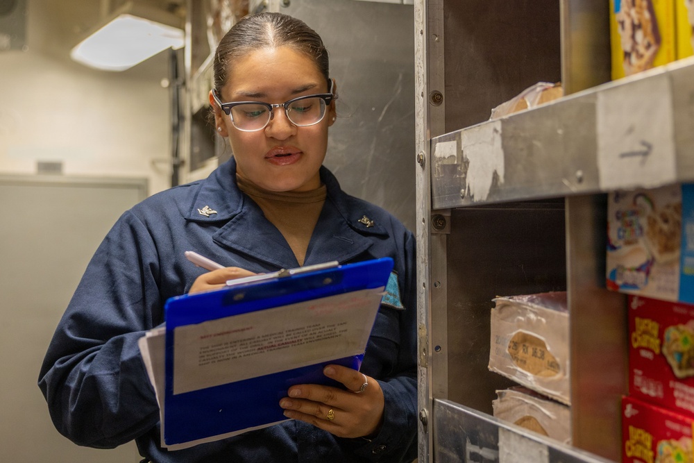A female military service member in a navy blue uniform is conducting an inventory check in what appears to be a ship or military installation's food storage area. She is holding a blue clipboard and writing or reviewing documents while standing next to metal shelving stocked with food items. Her uniform displays insignia consistent with a junior enlisted rank.