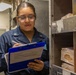 A female military service member in a navy blue uniform is conducting an inventory check in what appears to be a ship or military installation's food storage area. She is holding a blue clipboard and writing or reviewing documents while standing next to metal shelving stocked with food items. Her uniform displays insignia consistent with a junior enlisted rank.