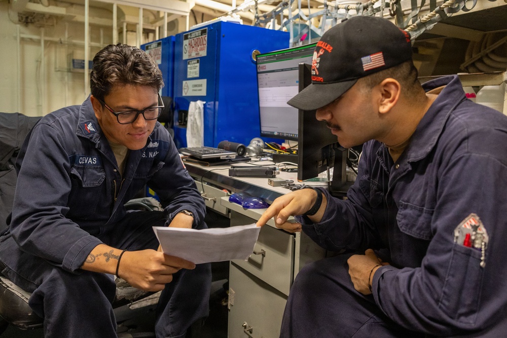 Two U.S. Navy sailors in dark blue coveralls are reviewing a document together in what appears to be a shipboard workspace or maintenance area. The sailor on the left, identified by a name tag reading 'CEVAS,' is holding a paper while the sailor on the right, wearing a black cap with an American flag patch, points to something on the document. A computer monitor, tools, and blue storage cabinets labeled 'HAZARDOUS' are visible in the background.