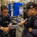 Two U.S. Navy sailors in dark blue coveralls are reviewing a document together in what appears to be a shipboard workspace or maintenance area. The sailor on the left, identified by a name tag reading 'CEVAS,' is holding a paper while the sailor on the right, wearing a black cap with an American flag patch, points to something on the document. A computer monitor, tools, and blue storage cabinets labeled 'HAZARDOUS' are visible in the background.