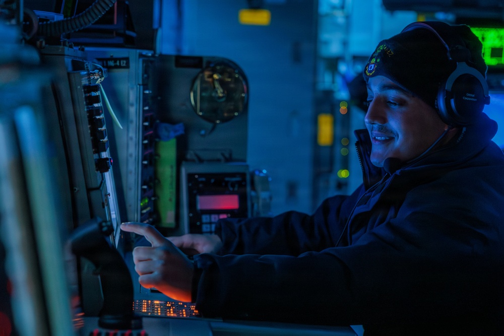 A military personnel member is seated at a control station in what appears to be a naval vessel's combat information center or bridge, illuminated by blue ambient lighting. The individual is wearing a dark beanie cap with an insignia, headphones, and a dark jacket, and is pointing at or operating a touchscreen display while smiling. The environment is filled with electronic equipment, control panels, and displays typical of a military operations room.