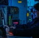 A military personnel member is seated at a control station in what appears to be a naval vessel's combat information center or bridge, illuminated by blue ambient lighting. The individual is wearing a dark beanie cap with an insignia, headphones, and a dark jacket, and is pointing at or operating a touchscreen display while smiling. The environment is filled with electronic equipment, control panels, and displays typical of a military operations room.