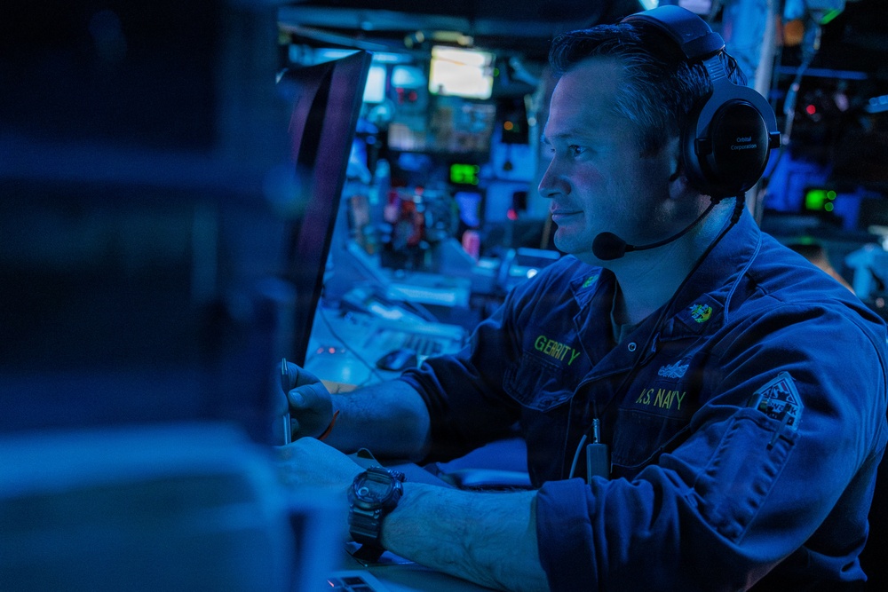 A U.S. Navy sailor identified by name tape as 'GERRITY' is seated at a combat or operations control station aboard a naval vessel. The environment is dimly lit with blue ambient lighting, characteristic of a Combat Information Center (CIC) or similar tactical operations room. The sailor is wearing a headset with boom microphone and appears to be actively monitoring or operating equipment.