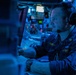 A U.S. Navy sailor identified by name tape as 'GERRITY' is seated at a combat or operations control station aboard a naval vessel. The environment is dimly lit with blue ambient lighting, characteristic of a Combat Information Center (CIC) or similar tactical operations room. The sailor is wearing a headset with boom microphone and appears to be actively monitoring or operating equipment.