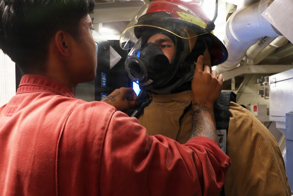 A person in a red shirt is assisting another individual in donning firefighting gear, including a helmet with a face shield and a breathing apparatus mask. The setting appears to be an interior space with visible pipes and equipment, consistent with a naval vessel or industrial facility. This appears to be a training or preparation exercise for emergency response.