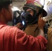 A person in a red shirt is assisting another individual in donning firefighting gear, including a helmet with a face shield and a breathing apparatus mask. The setting appears to be an interior space with visible pipes and equipment, consistent with a naval vessel or industrial facility. This appears to be a training or preparation exercise for emergency response.