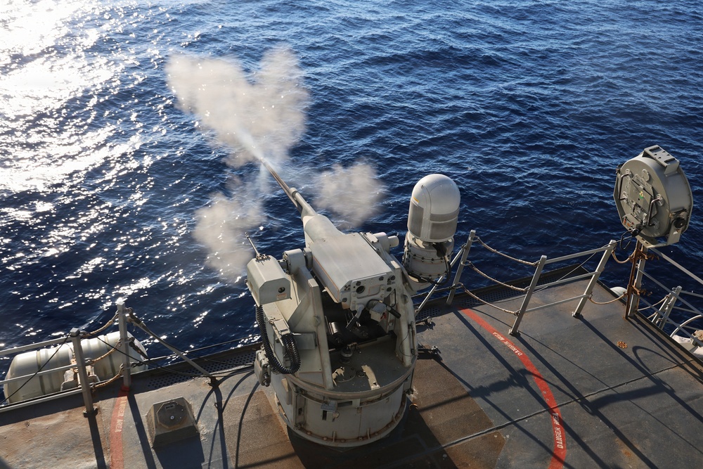 A naval deck-mounted weapon system is actively firing, with visible muzzle smoke/blast discharge extending toward the open ocean. The image is taken from an elevated angle on what appears to be a military vessel's deck. The scene captures a live-fire exercise or operational engagement at sea.