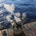 A naval deck-mounted weapon system is actively firing, with visible muzzle smoke/blast discharge extending toward the open ocean. The image is taken from an elevated angle on what appears to be a military vessel's deck. The scene captures a live-fire exercise or operational engagement at sea.