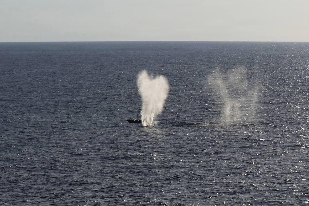 The image shows an open ocean scene with what appears to be a submarine partially surfacing or submerging, accompanied by two distinct water spout or blow formations rising from the sea surface. The larger central spout has a broad, mushroom-like shape while a second narrower column of mist or spray is visible to the right. The scene is photographed from an elevated vantage point over calm, dark blue ocean water.