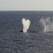 The image shows an open ocean scene with what appears to be a submarine partially surfacing or submerging, accompanied by two distinct water spout or blow formations rising from the sea surface. The larger central spout has a broad, mushroom-like shape while a second narrower column of mist or spray is visible to the right. The scene is photographed from an elevated vantage point over calm, dark blue ocean water.