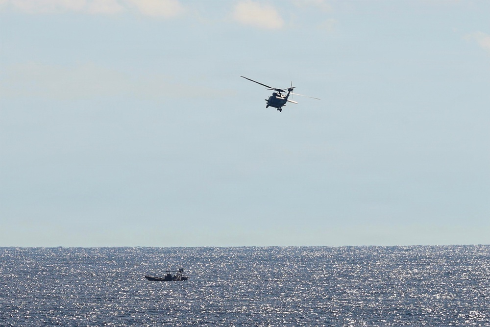 The image shows a military or coast guard helicopter flying over open ocean water, with a small vessel visible on the water's surface below. The scene appears to depict a maritime patrol or search-and-rescue operation. The photograph is taken from a distance, capturing both the airborne and surface assets against a partly cloudy sky.