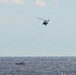 The image shows a military or coast guard helicopter flying over open ocean water, with a small vessel visible on the water's surface below. The scene appears to depict a maritime patrol or search-and-rescue operation. The photograph is taken from a distance, capturing both the airborne and surface assets against a partly cloudy sky.
