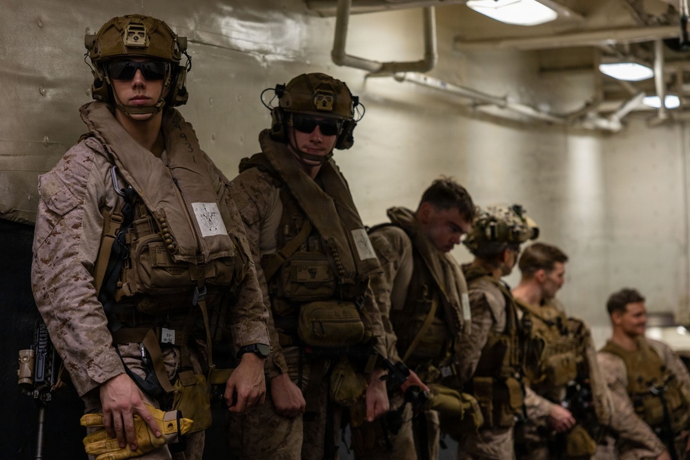 A group of military personnel, appearing to be U.S. Marines based on their MARPAT desert camouflage uniforms, are lined up in what appears to be the interior of a naval vessel or military facility. They are fully equipped with tactical gear, helmets with night-vision mounts, body armor, and weapons. The personnel appear to be staged and ready for a mission or operation.