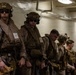 A group of military personnel, appearing to be U.S. Marines based on their MARPAT desert camouflage uniforms, are lined up in what appears to be the interior of a naval vessel or military facility. They are fully equipped with tactical gear, helmets with night-vision mounts, body armor, and weapons. The personnel appear to be staged and ready for a mission or operation.