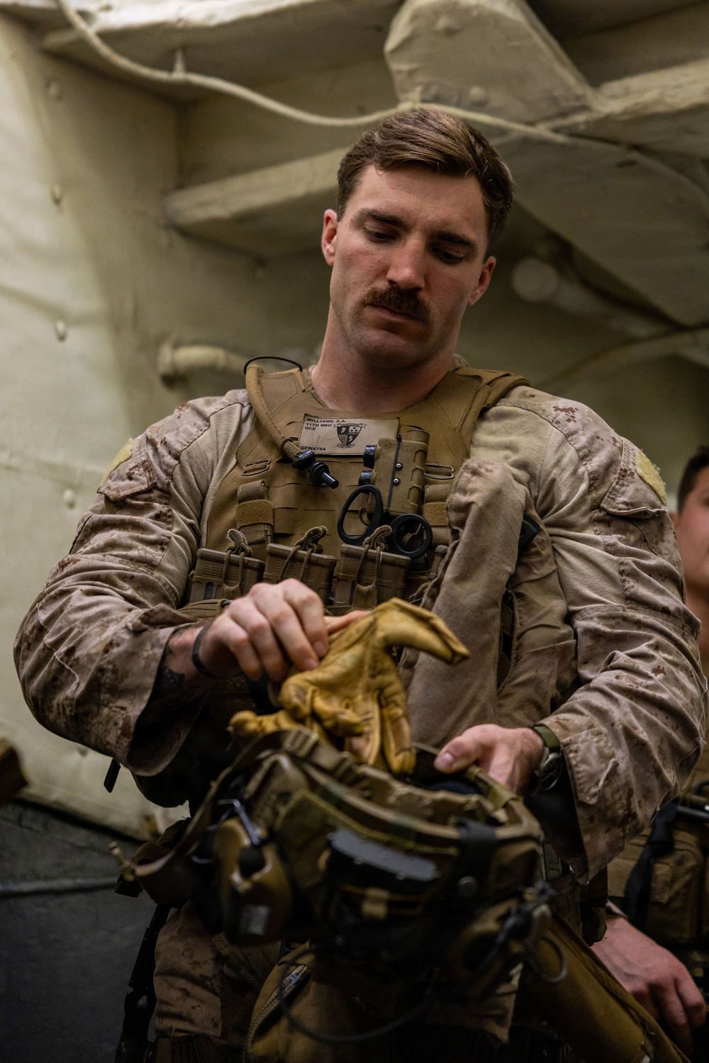 A male military service member in desert camouflage (MARPAT) uniform and tactical vest is preparing equipment, appearing to handle tan tactical gloves. He is indoors in what appears to be a ship or military facility interior. A name tape and unit patch are visible on his chest rig.