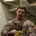 A male military service member in desert camouflage (MARPAT) uniform and tactical vest is preparing equipment, appearing to handle tan tactical gloves. He is indoors in what appears to be a ship or military facility interior. A name tape and unit patch are visible on his chest rig.