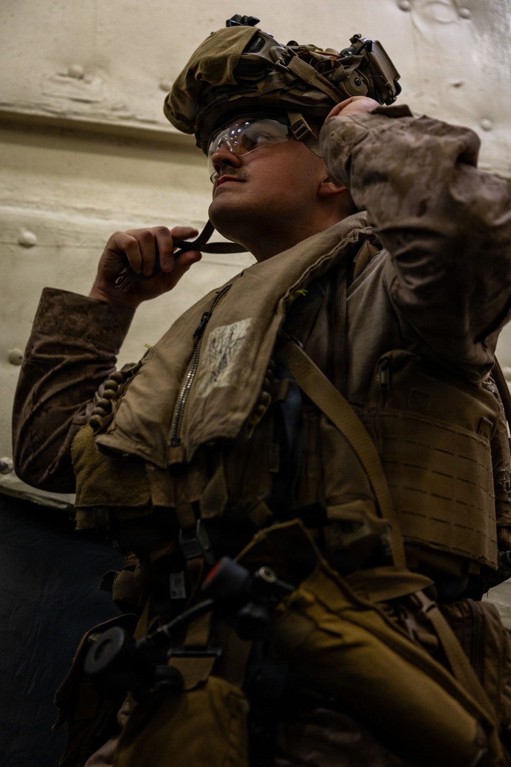 A heavily equipped military service member is seated against a light-colored metal wall, looking upward. The individual is wearing full combat gear including a tactical helmet with mounted accessories, body armor, and carrying a large pack. The low-angle photograph creates a dramatic perspective emphasizing the weight and complexity of the equipment.