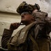 A heavily equipped military service member is seated against a light-colored metal wall, looking upward. The individual is wearing full combat gear including a tactical helmet with mounted accessories, body armor, and carrying a large pack. The low-angle photograph creates a dramatic perspective emphasizing the weight and complexity of the equipment.