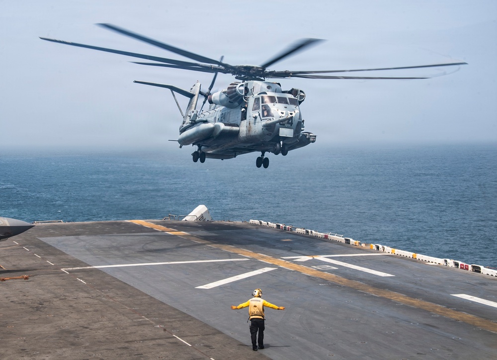 A large military helicopter, identifiable as a CH-53E Super Stallion, is hovering low over the flight deck of a naval vessel, likely an amphibious assault ship. A flight deck crew member wearing a yellow jersey and ear protection is standing on the deck with arms outstretched, signaling the helicopter during landing or departure operations. The ocean is visible in the background under an overcast sky.