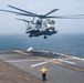 A large military helicopter, identifiable as a CH-53E Super Stallion, is hovering low over the flight deck of a naval vessel, likely an amphibious assault ship. A flight deck crew member wearing a yellow jersey and ear protection is standing on the deck with arms outstretched, signaling the helicopter during landing or departure operations. The ocean is visible in the background under an overcast sky.